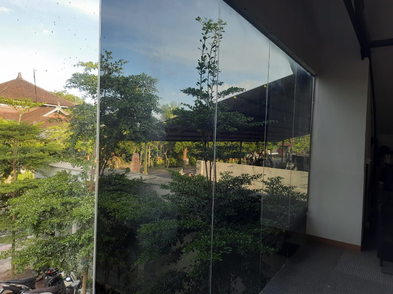 Dinding kaca dengan kaca film reflektif gelap yang memantulkan pepohonan dan atap bergaya Bali
